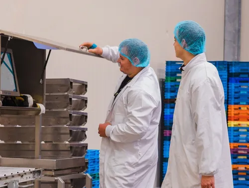 2 medewerkers met witte overjas en blauw mutsje op kijken naar een productregistratiestation